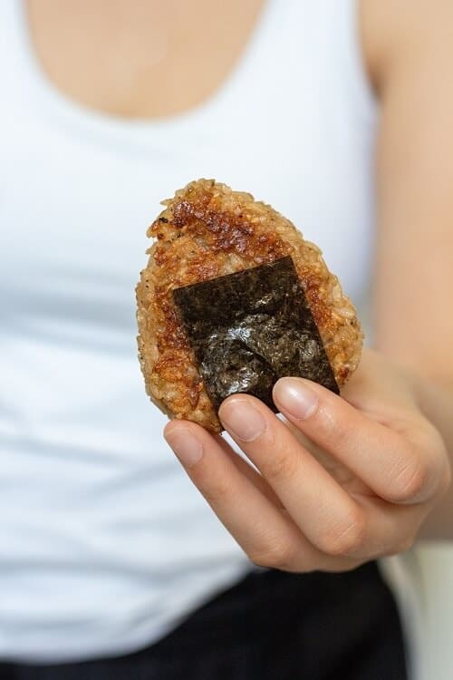 Person holding a triangle of golden-brown pan-fried rice wrapped with a piece of nori