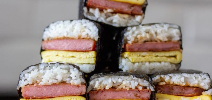 Stack of 6 spam musubi with egg on a wooden cutting board.