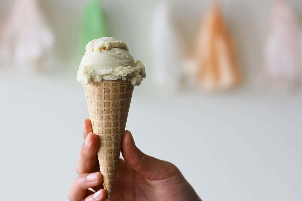 Hand holding a sugar cone with a scoop of beige ice cream on top.
