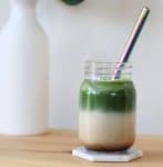 Clear glass jar filled with three layers: espresso, milk and matcha. There's a vase of flowers in the background.