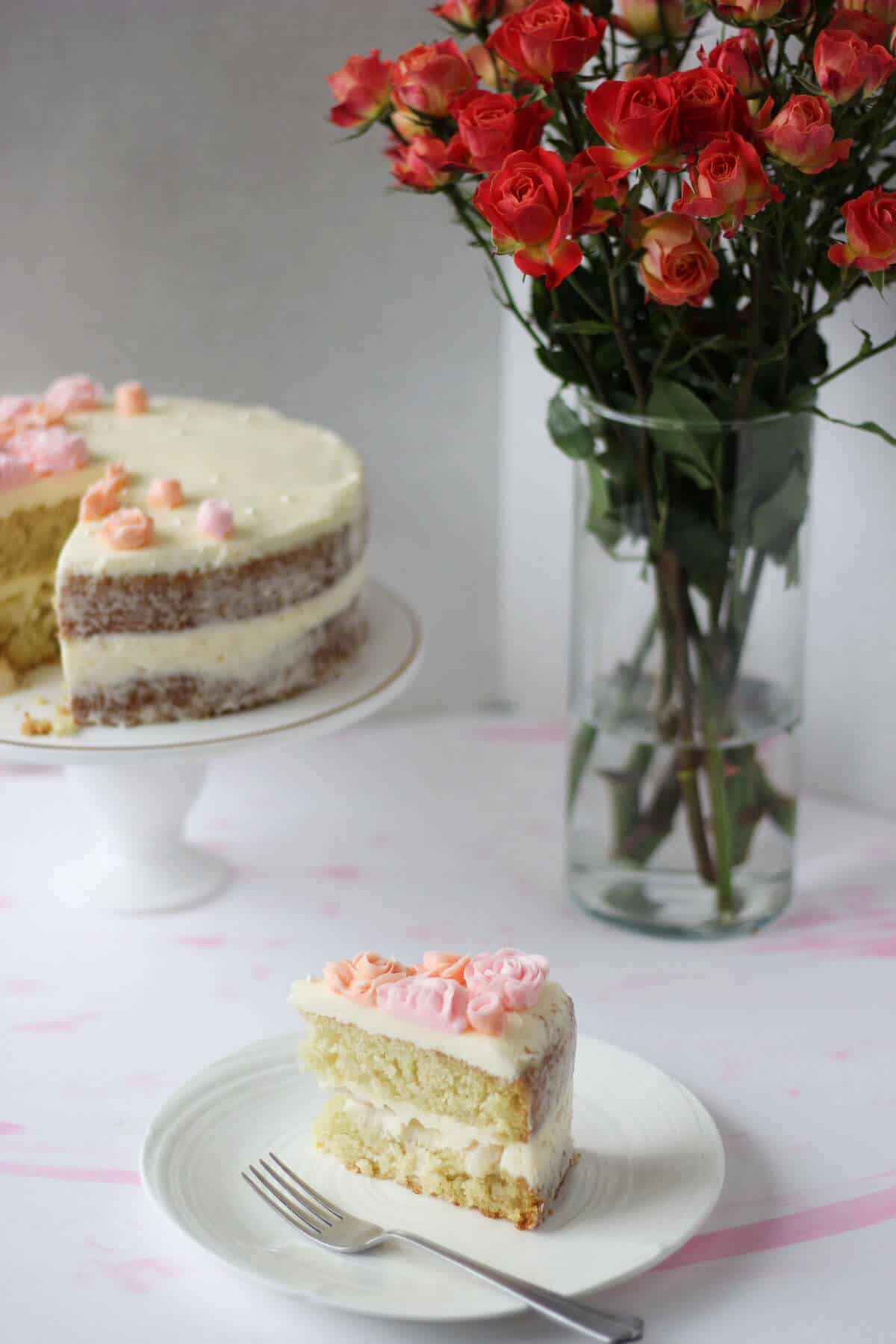 Slice of cake in the forefront with the rest of the cake and a vase of pink roses in the background.