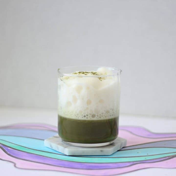 Cool-toned background and a marble coaster below a clear glass filled with a shot of bright green matcha and white milk foam.
