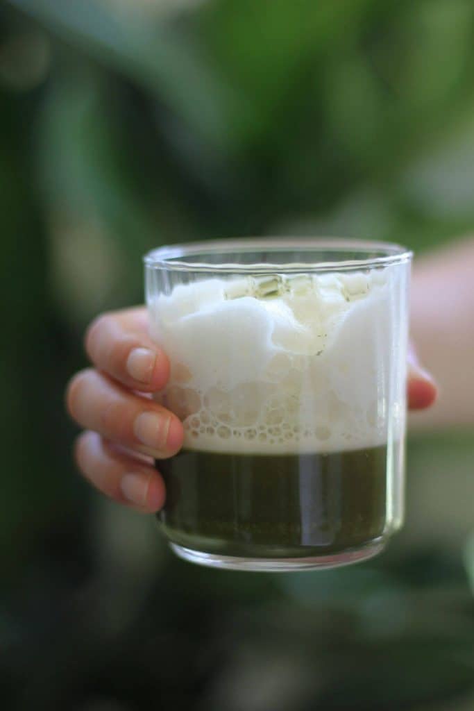 Hand holding clear glass with a shot of matcha at the bottom, topped with white milk foam.