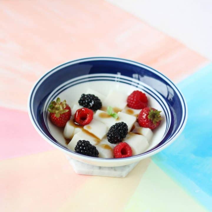 White bowl filled with white cubes of almond tofu, berries and brown sugar syrup.