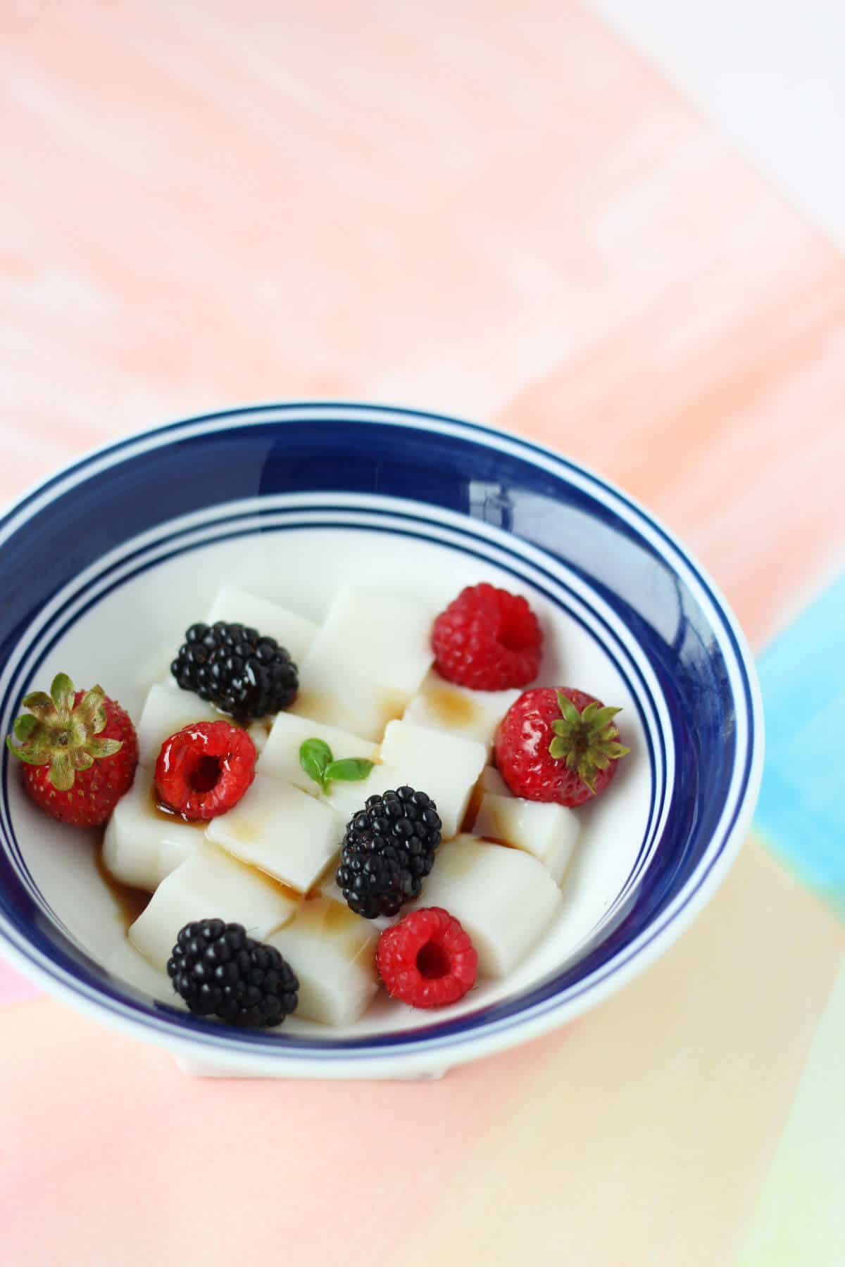 White bowl with blue rim, filled with cubes of white opaque almond tofu and topped with summer berries and brown sugar syrup.