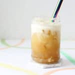 White coaster with clear glass filled with orange colored tea, ice, white cheese foam and a metal straw.