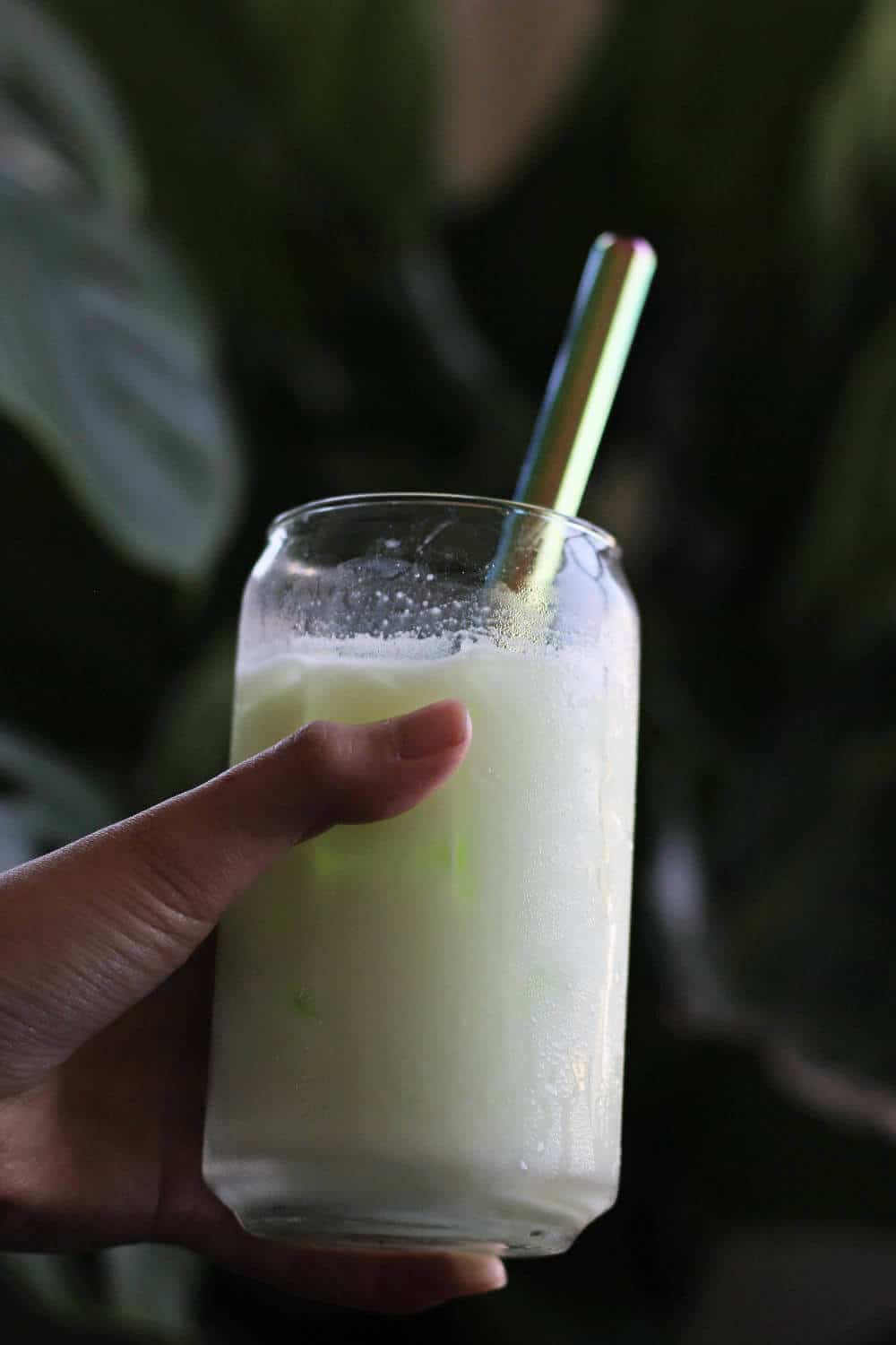 Hand holding clear cup filled with opaque, slightly green liquid and a metal straw.