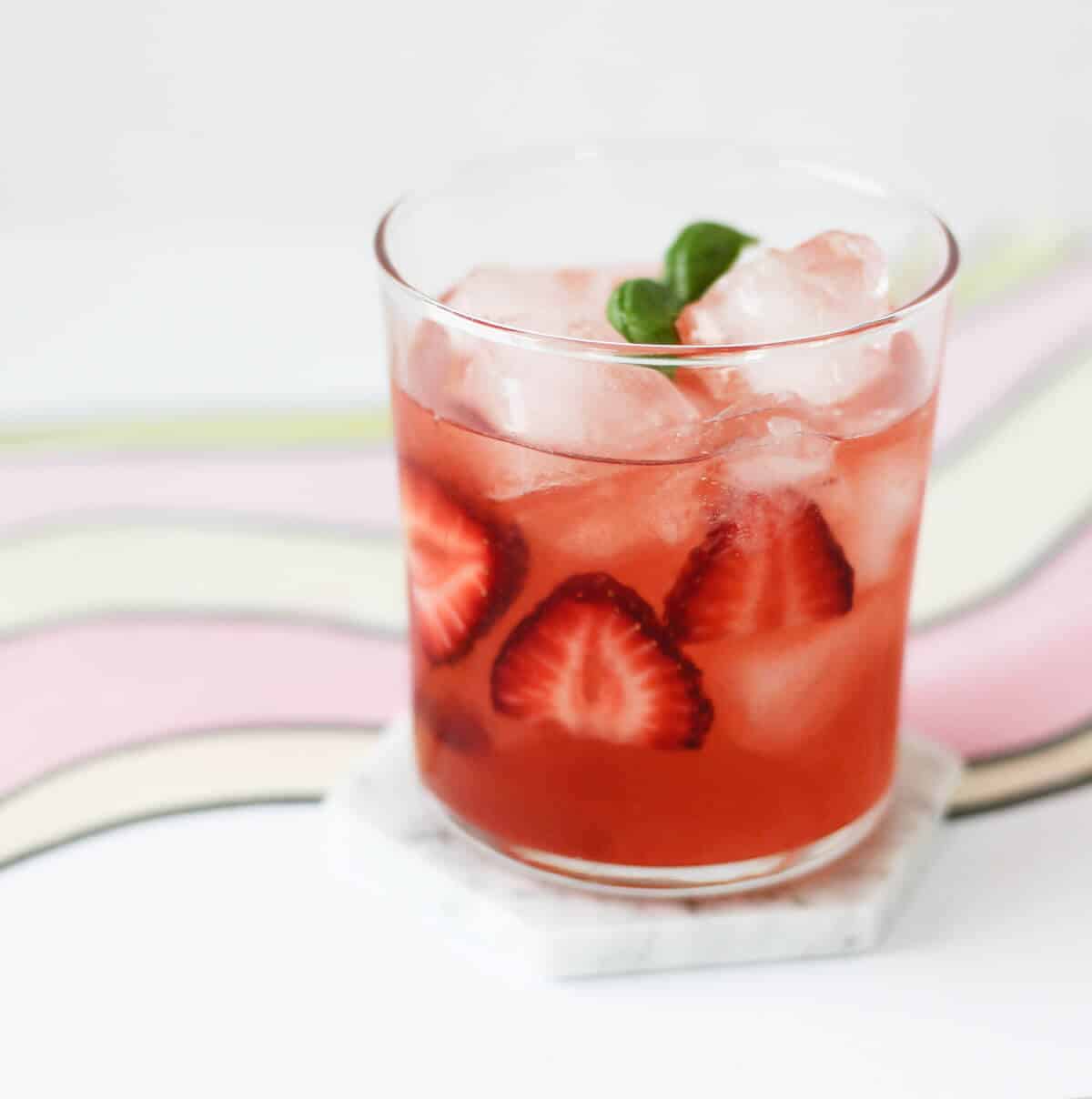 Clear glass filled with pink cocktail, sliced strawberries, ice and a sprig of basil.