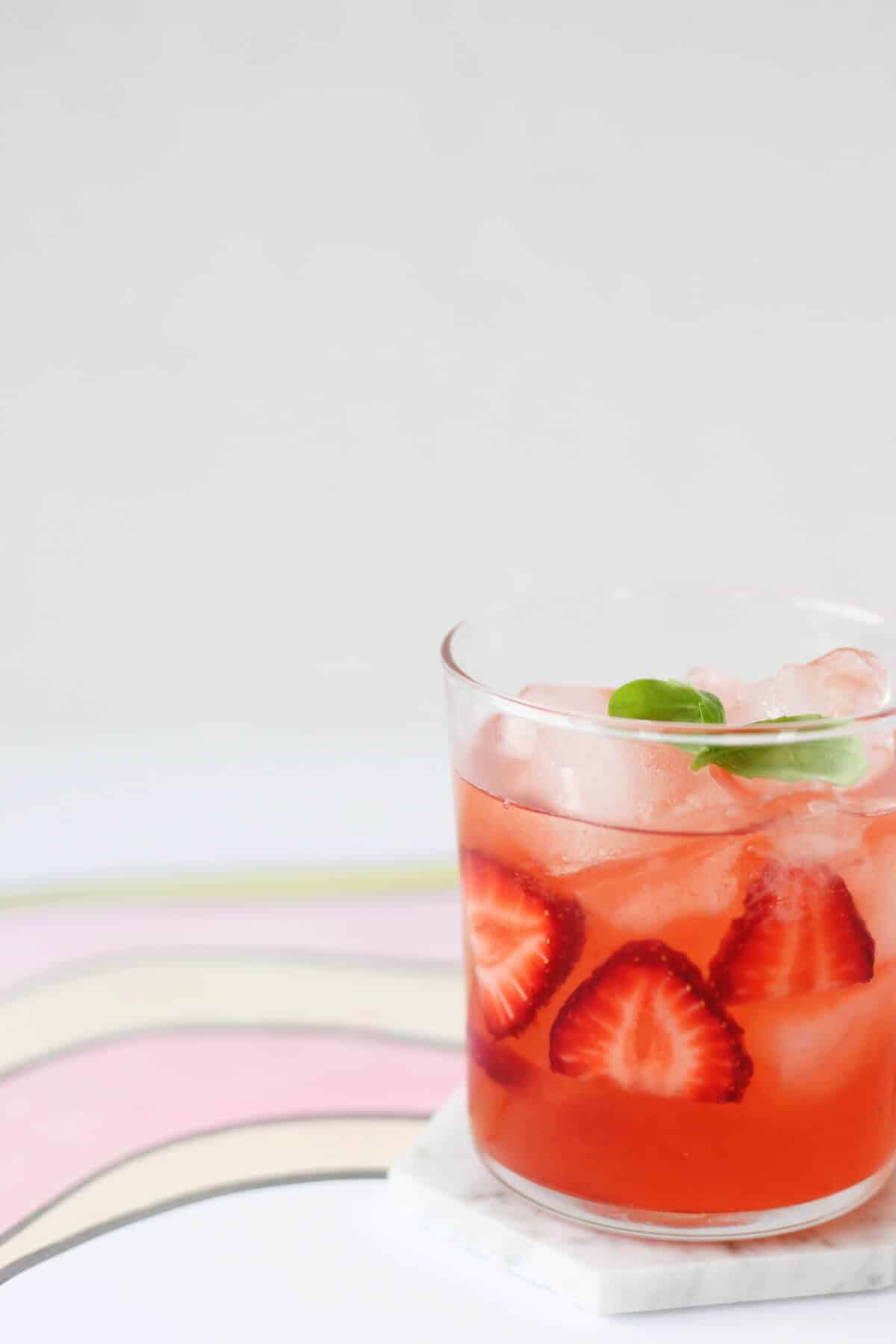 Clear glass filled with pink cocktail, sliced strawberries, ice and a sprig of basil.