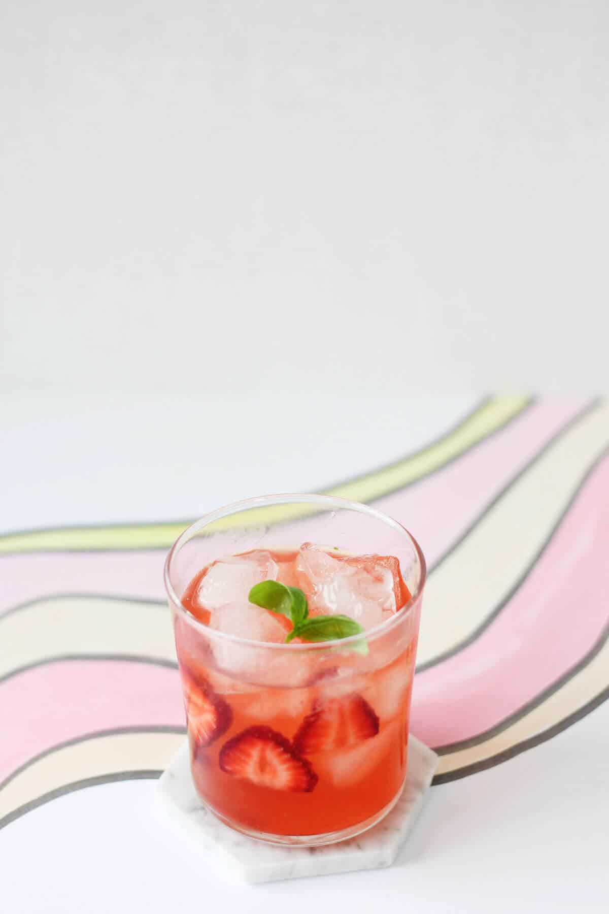 Clear glass filled with pink cocktail, sliced strawberries, ice and a sprig of basil.