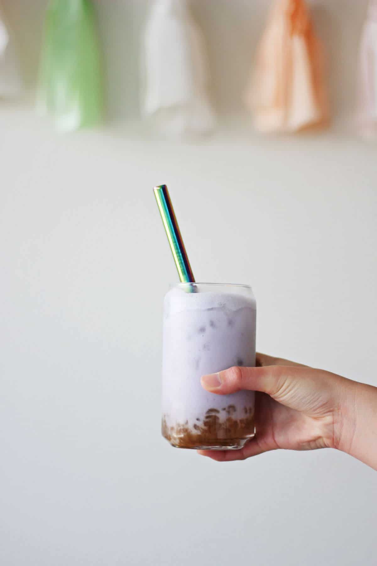 Hand holding clear glass filled with brown sugar boba, ice, lilac colored milk tea and a metal straw.