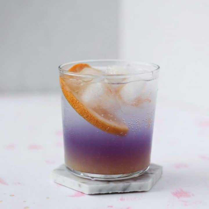 Clear glass filled with grapefruit juice, purple gin, a slice of grapefruit and tonic on top.