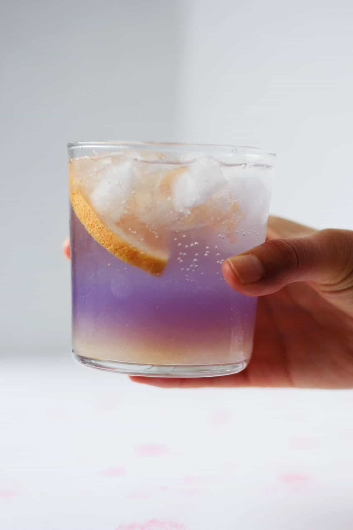 Hand holding clear glass filled with layered grapefruit juice and purple gin, topped with a slice of grapefruit and ice.