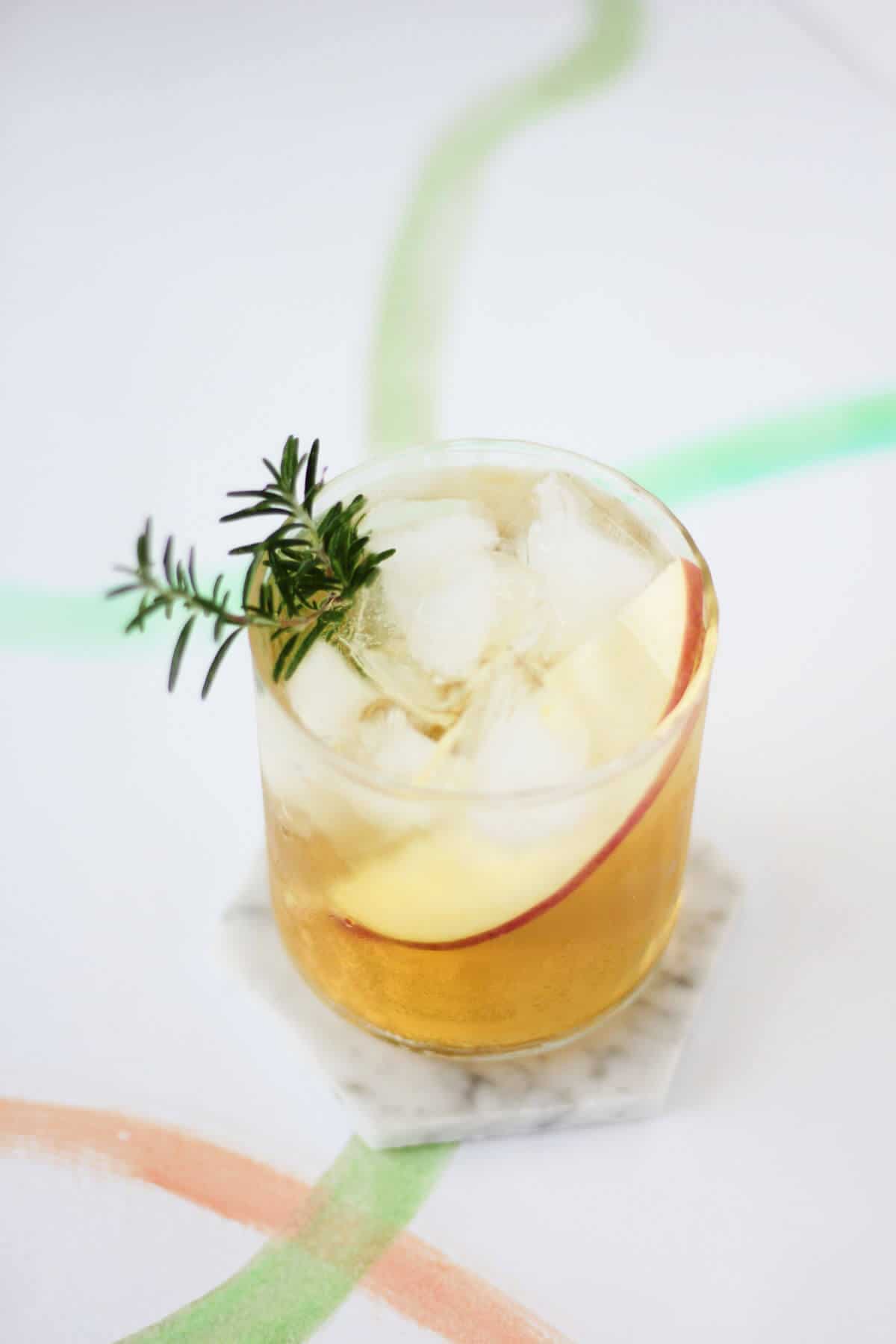 Clear glass filled with golden yellow liquid, a slice of apple and a sprig of rosemary.
