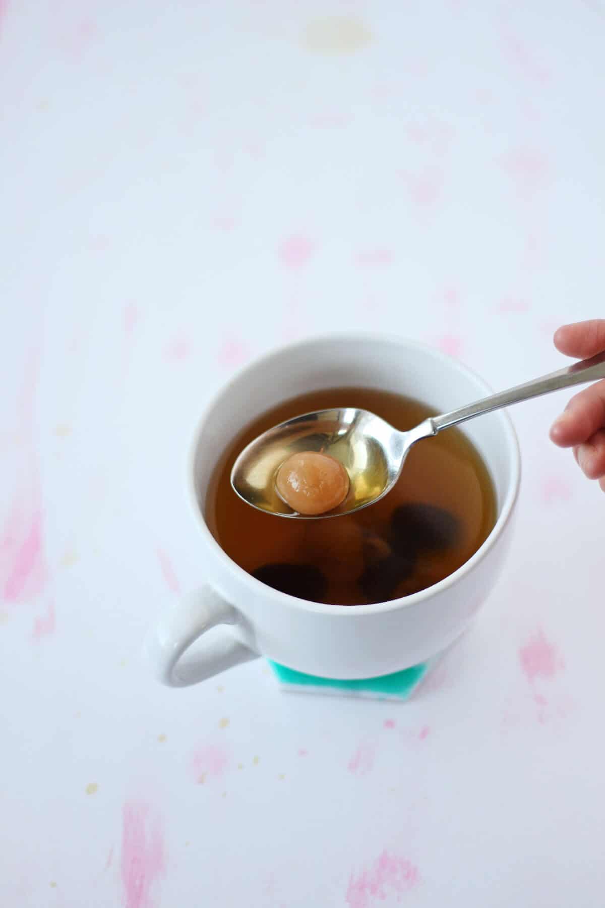  Hand holding spoon filled with longan over cup of reddish brown tea.