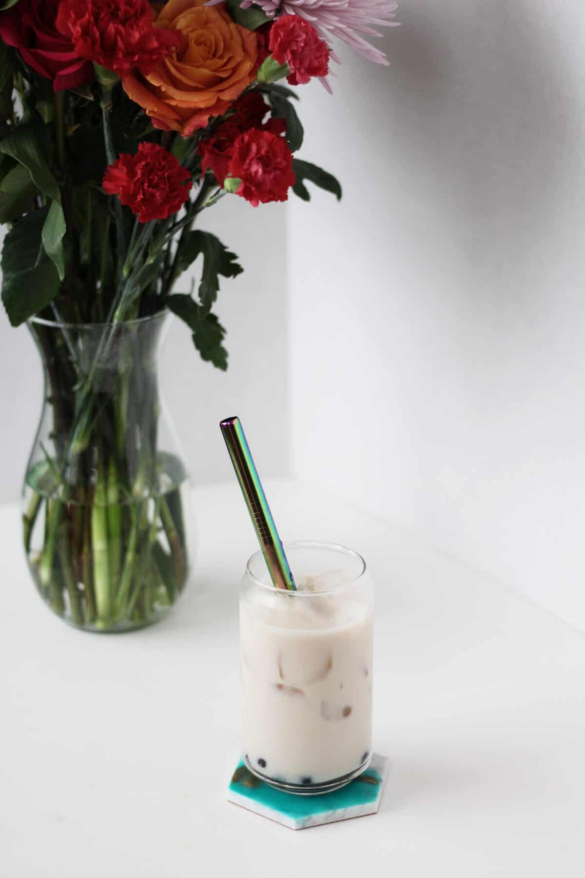 Clear glass cup filled with honey boba, ice and off white colored oolong milk tea with a metal straw. Vase of red flowers in the background.