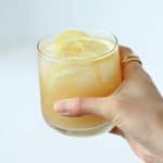 Hand holding a glass of orange and yellow hued liquid with thin slices of lemon on top.