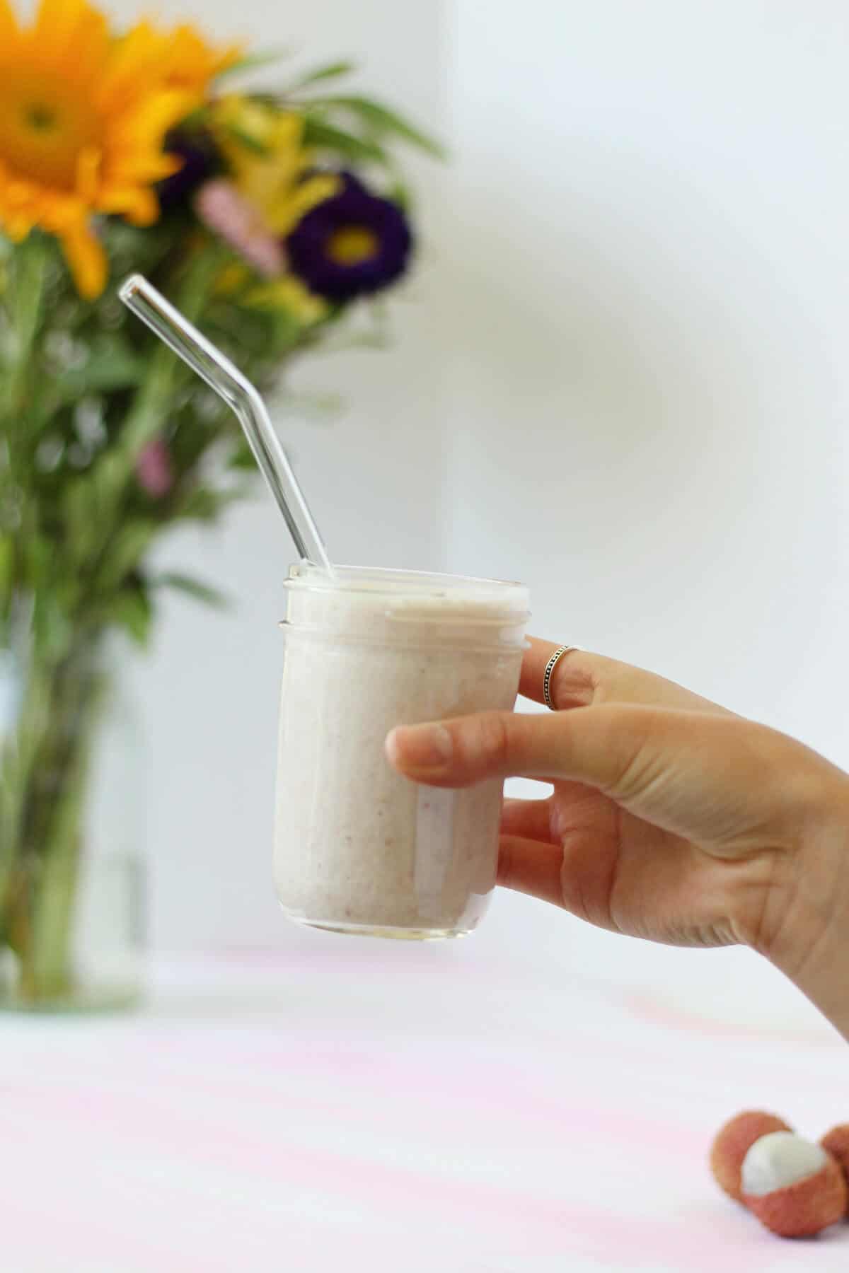 Hand holding a glass jar with a light pink lychee smoothie and a glass straw in it.