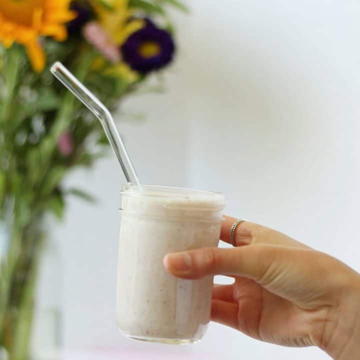Hand holding a glass jar with a light pink lychee smoothie and a glass straw in it.
