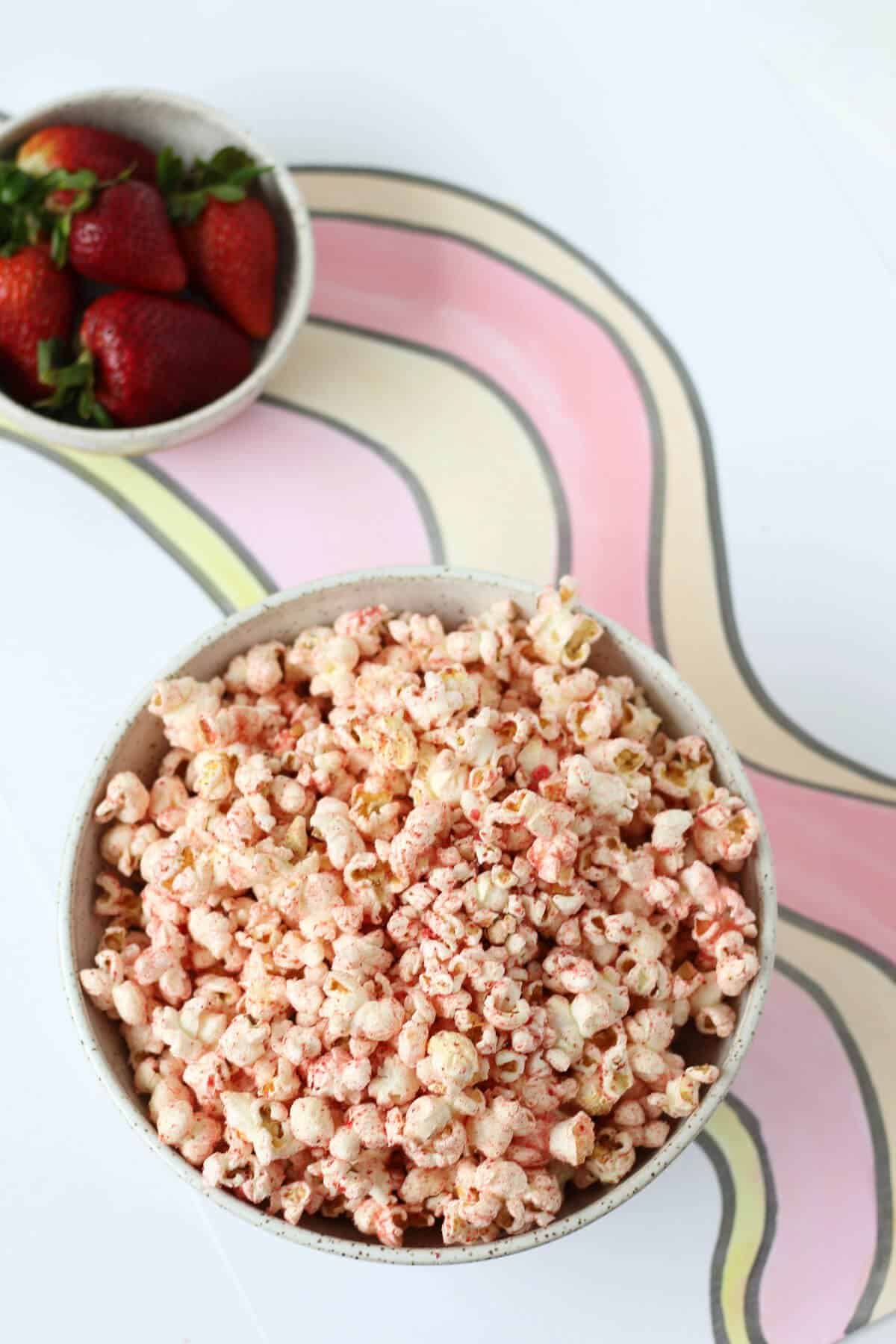 Bowl of strawberry sugar coated popcorn with a bowl of fresh strawberries in the background.