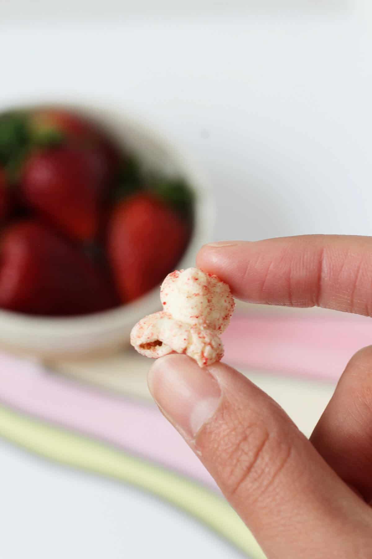 Fingers holding a piece of pink popcorn with a bowl of strawberries in the background.