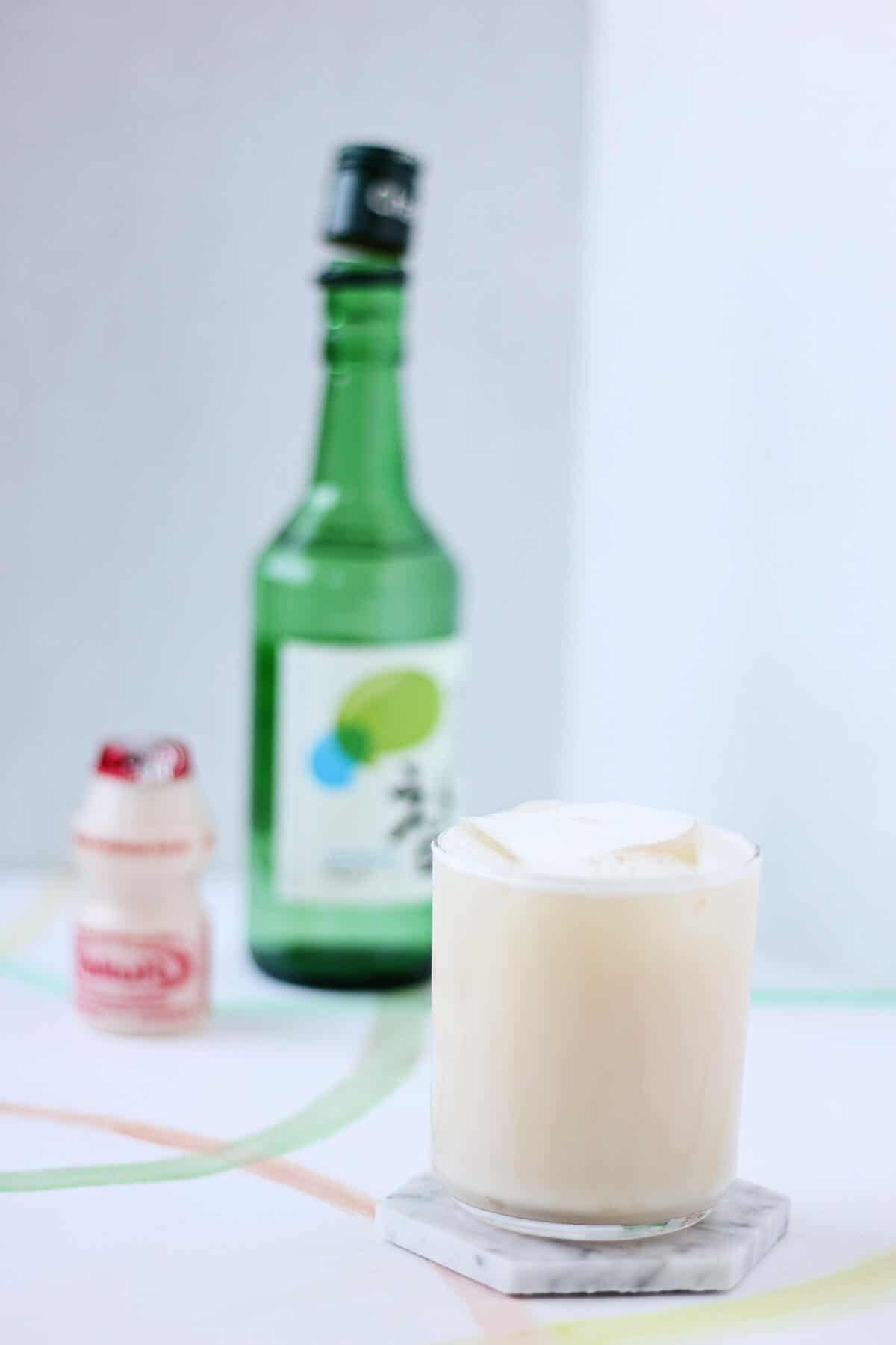 Clear glass of creamy off white liquid and ice in forefront on white marble coaster with a bottle of soju and a single serving container of Yakult in the back.