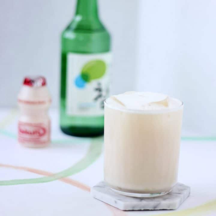 Clear glass of creamy off white liquid and ice in forefront on white marble coaster with a bottle of soju and a single serving container of Yakult in the back.