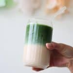 Hand holding clear glass filled with ice, pale orange Thai tea and bright green matcha.