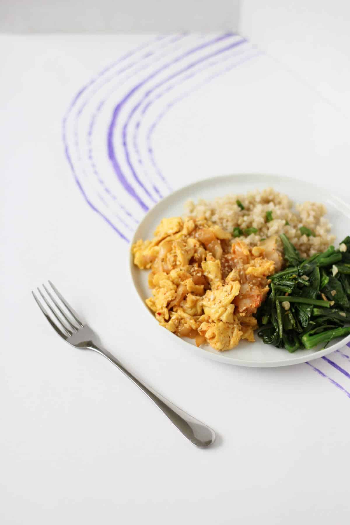 White plate with steamed rice, kimchi and scrambled eggs, and sauteed greens.