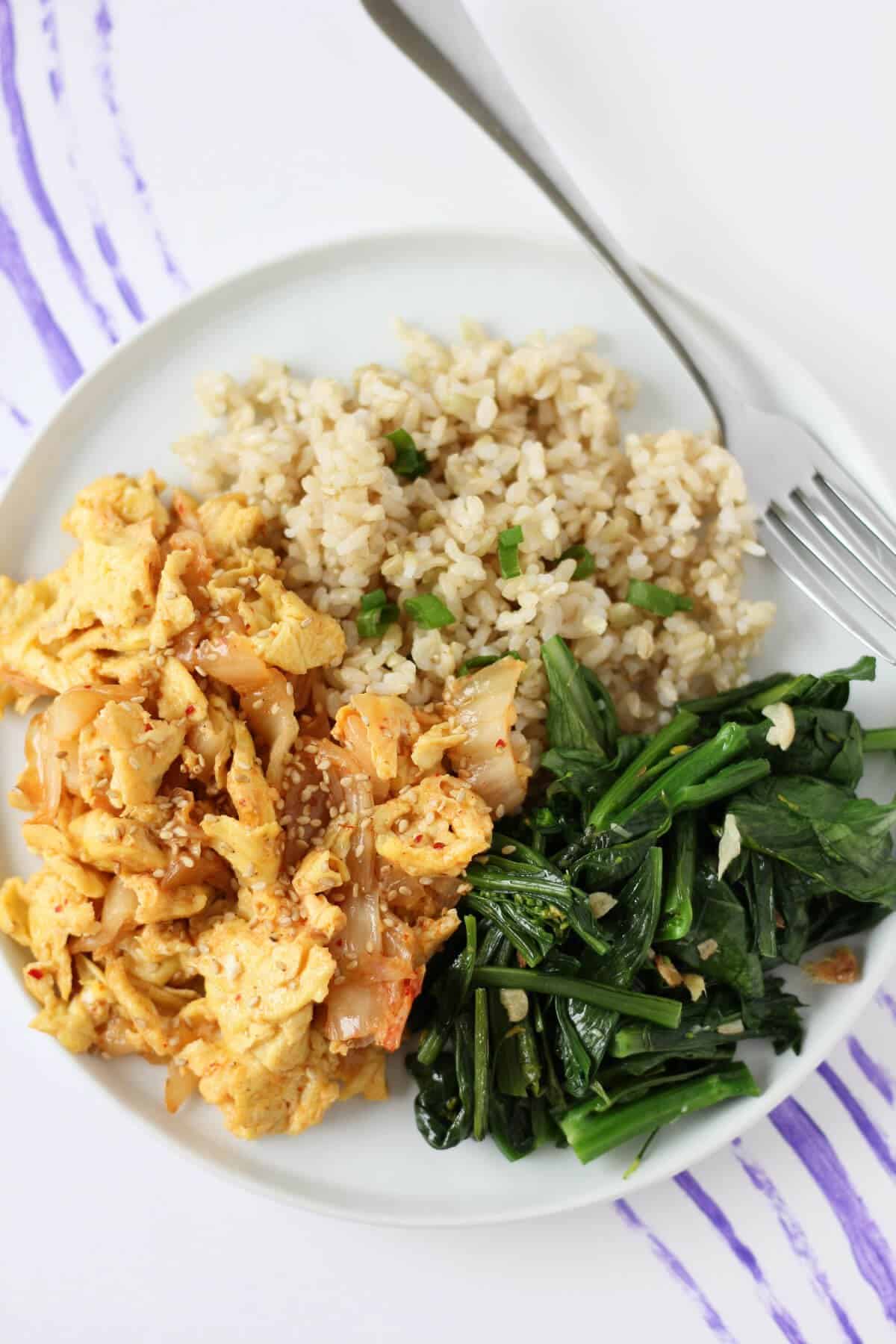 White plate with steamed rice, kimchi and scrambled eggs, and sauteed greens.