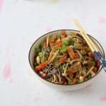 Bowl filled with colorful vegetables, pan-fried tofu and brown soba. There's a pair of chopsticks perched on the top of the bowl.