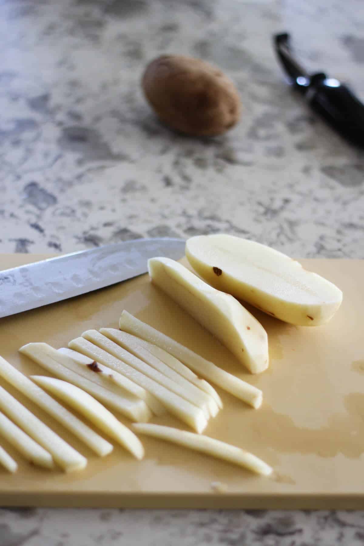 A cutting board with potatoes cut into fries with a whole potato and a vegetable peeler in the background.