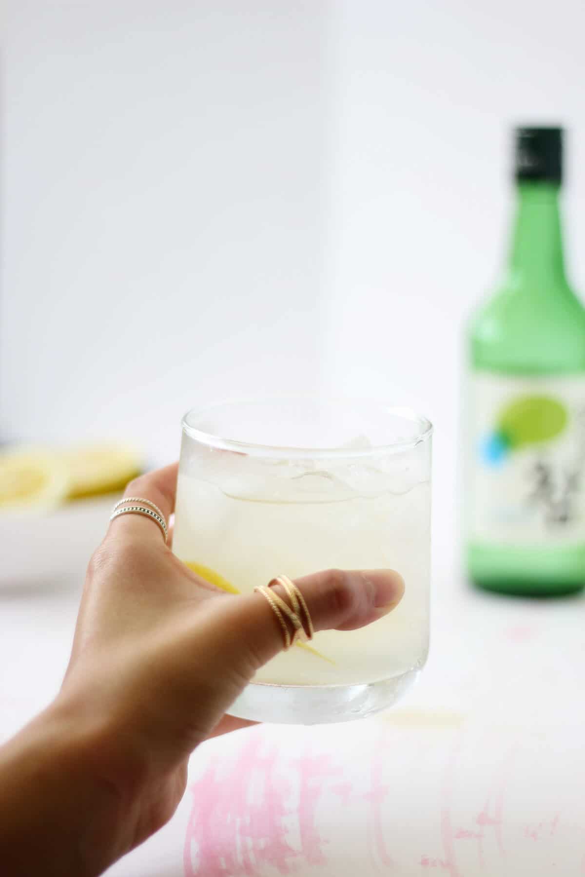 Hand with a thumb ring holding a clear glass filled with light yellow liquid. There's a bowl of lemons and a bottle of soju in the background.
