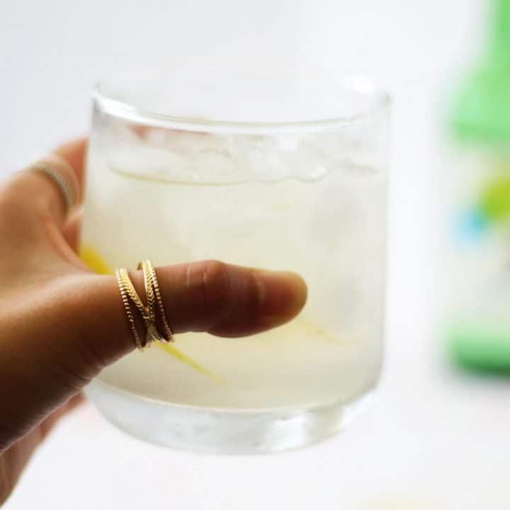 Hand with a thumb ring holding a clear glass filled with light yellow liquid.