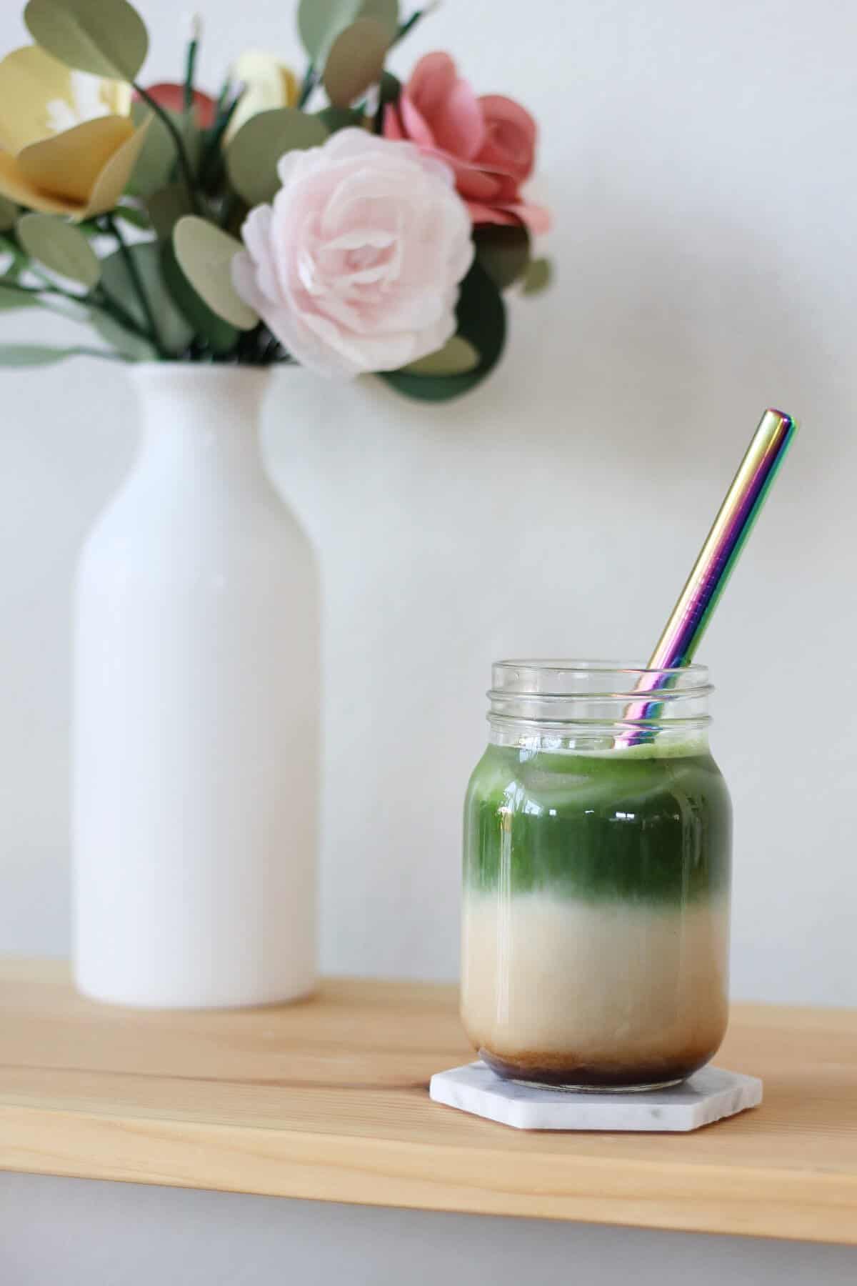 Clear glass jar filled with three layers: espresso, milk and matcha. There's a vase of flowers in the background.