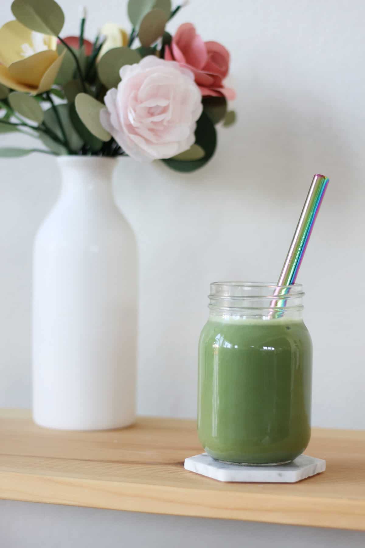 Clear jar with bright green matcha and a straw. There's a vase of flowers in the background.