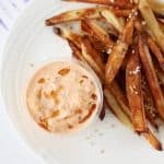 A little container filled with creamy orange-tinted kimchi mayo topped with a drizzle of sesame oil. There is a side of fries.