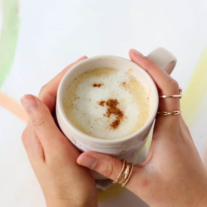 Hands holding a mug of coffee with steamed milk and cinnamon on top.