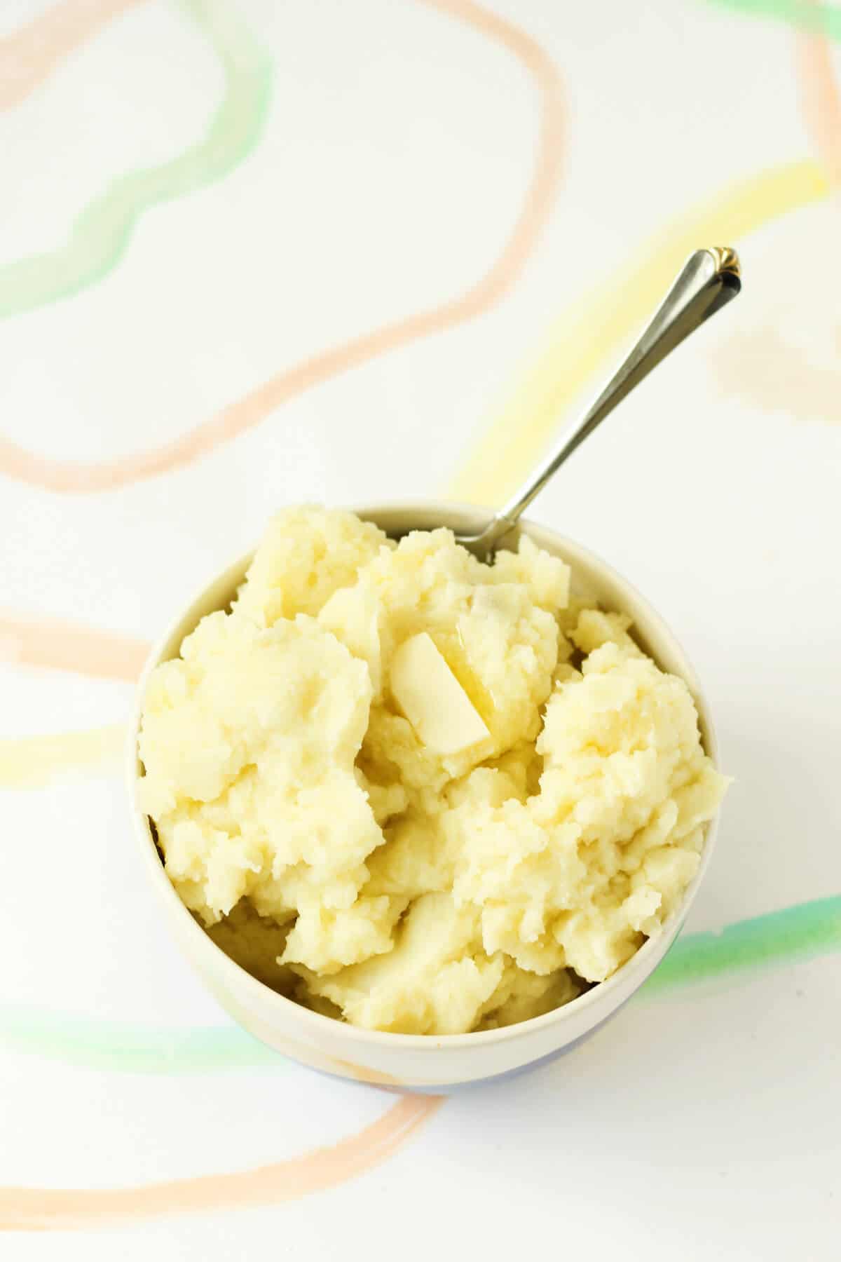 White bowl filled with yellow-y mashed potatoes topped with a pat of melty butter. There's a metal spoon sticking out of the bowl.