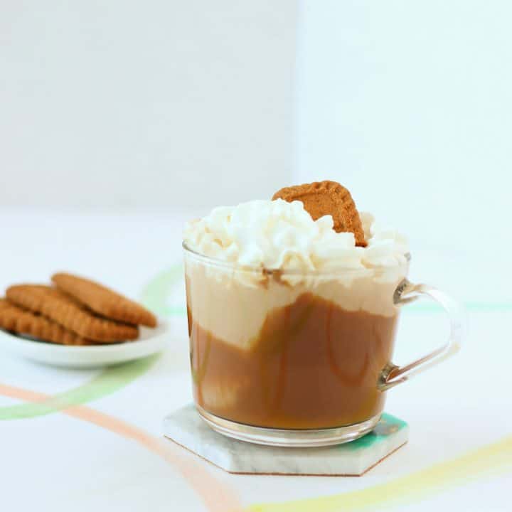 Clear mug smeared with brown cookie butter, then filled with tan coffee, and topped with whipped cream and a cookie. There's a little dish of cookies in the background.