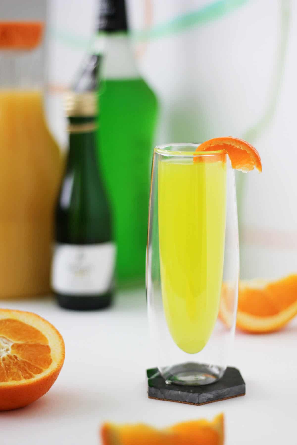 Tall clear glass with insulated walls with a lemon-lime colored liquid, garnished with a slice of tangerine on the rim. There are orange slices, as well as the juice, champagne and liqueur bottles, in the background.