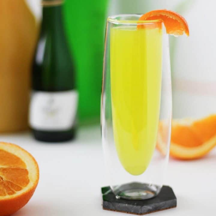 Tall clear glass with insulated walls with a lemon-lime colored liquid, garnished with a slice of tangerine on the rim. There are orange slices, as well as the juice, champagne and liqueur bottles, in the background.