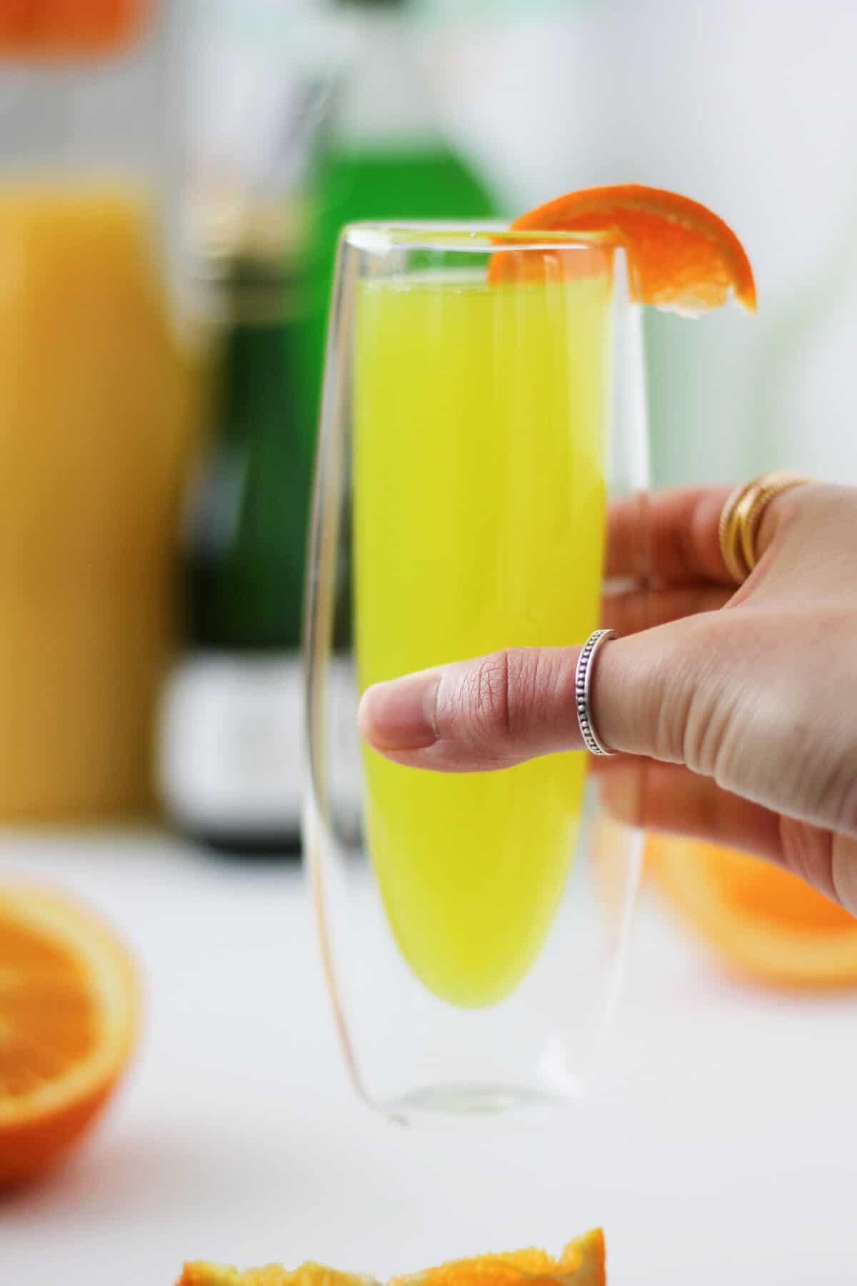 Hand holding a tall clear glass with insulated walls with a lemon-lime colored liquid, garnished with a slice of tangerine on the rim. There are orange slices, as well as the juice, champagne and liqueur bottles, in the background.