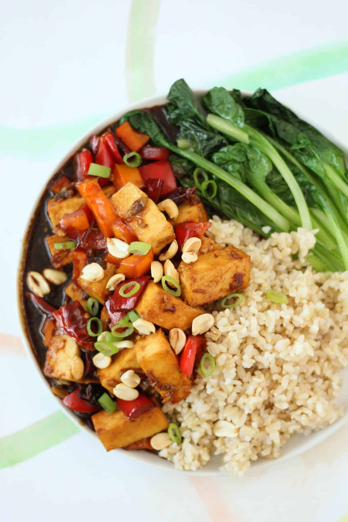Plate filled with chunks of paneer in a reddish brown gravy, mixed with red bell peppers, golden peanuts and sliced green onions. The rest of the plate is filled with steamed green leafy vegetables and steamed brown rice.