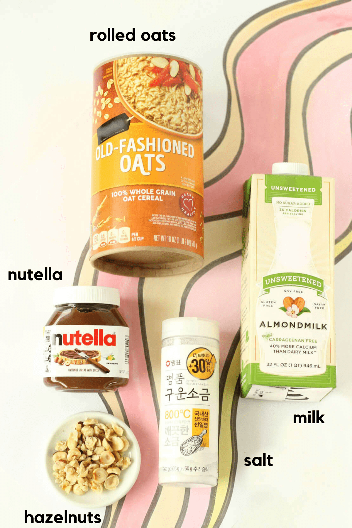Ingredients laid out for Nutella overnight oats, including a canister of rolled oats, a carton of soy milk, salt, a jar of Nutella and chopped toasted hazelnuts.