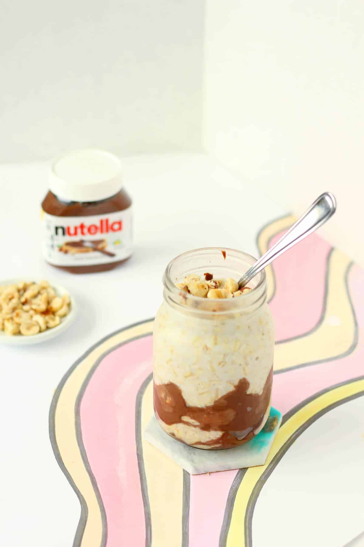 A clear glass jar with Nutella smeared on the inside with cream-colored overnight oats and topped with chopped hazelnuts. There's a spoon sticking out of the jar, with a bowl of hazelnuts and a jar of Nutella in the background.