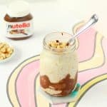 A clear glass jar with Nutella smeared on the inside with cream-colored overnight oats and topped with chopped hazelnuts. There's a spoon sticking out of the jar, with a bowl of hazelnuts and a jar of Nutella in the background.