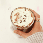 Hand with rings on the second finger holding a clear mug of hot chocolate topped with whipped cream and chocolate shavings.