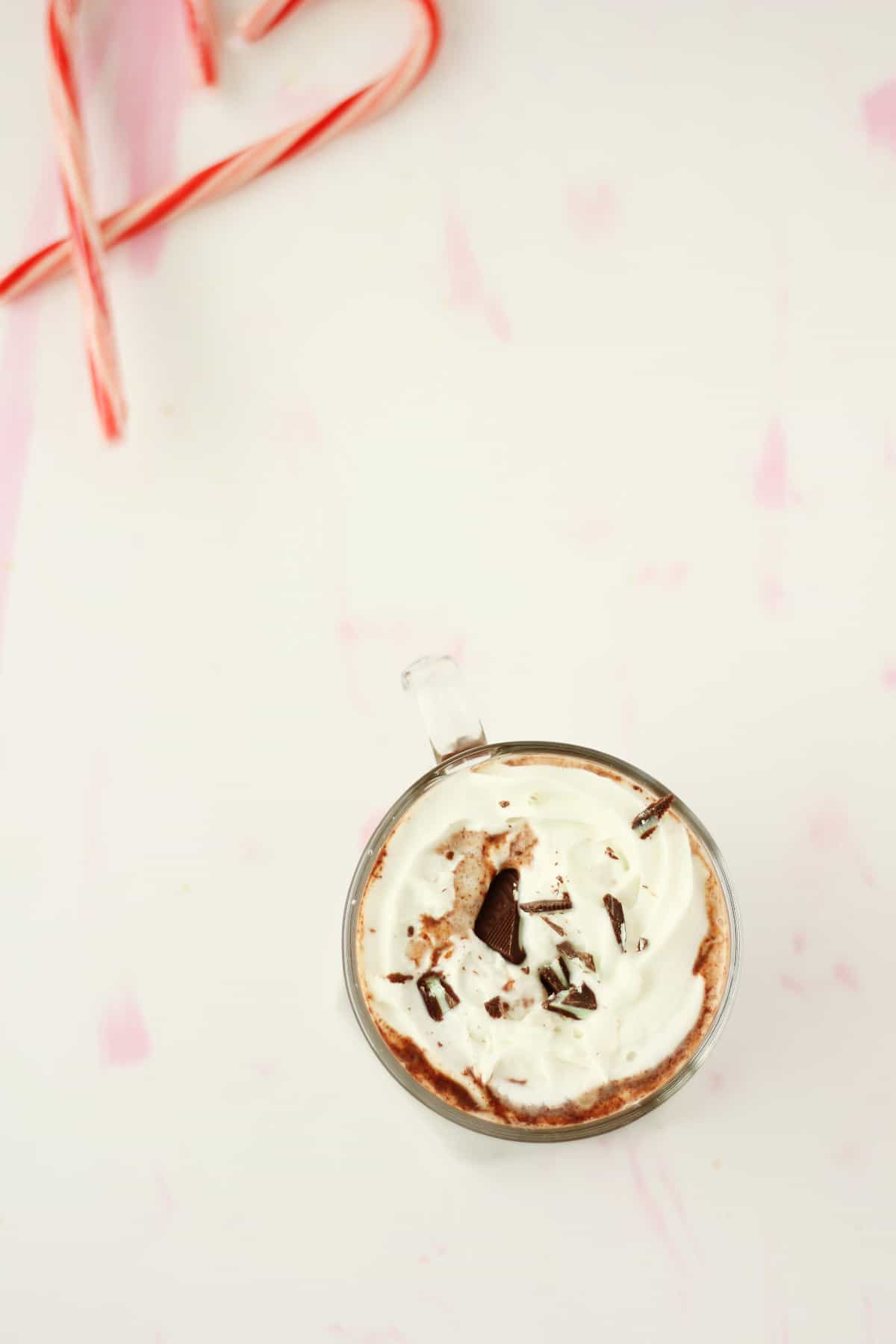 Overhead view of a clear mug filled with hot chocolate, whipped cream and chocolate shavings. There are a few candy canes blurred in the background.