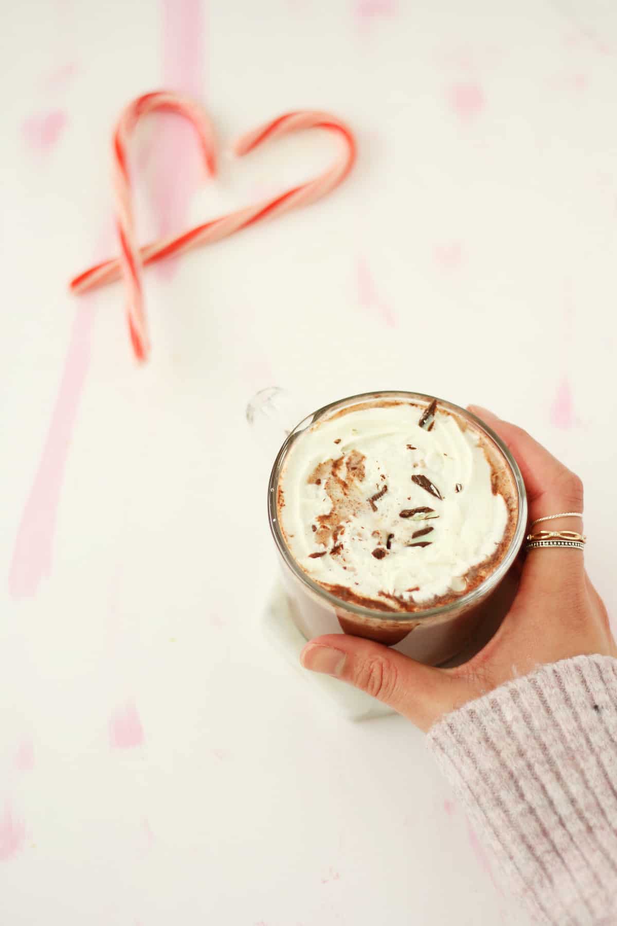 Hand with rings on the second finger holding a clear mug of hot chocolate topped with whipped cream and chocolate shavings. There are a few candy canes in the background.
